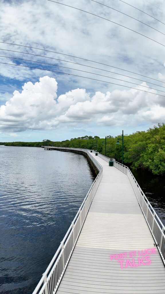 A long white boardwalk curving along the North Fork of the Saint Lucie River in Port Saint Lucie, Florida, surrounded by calm water and lush green mangroves under a partly cloudy sky.